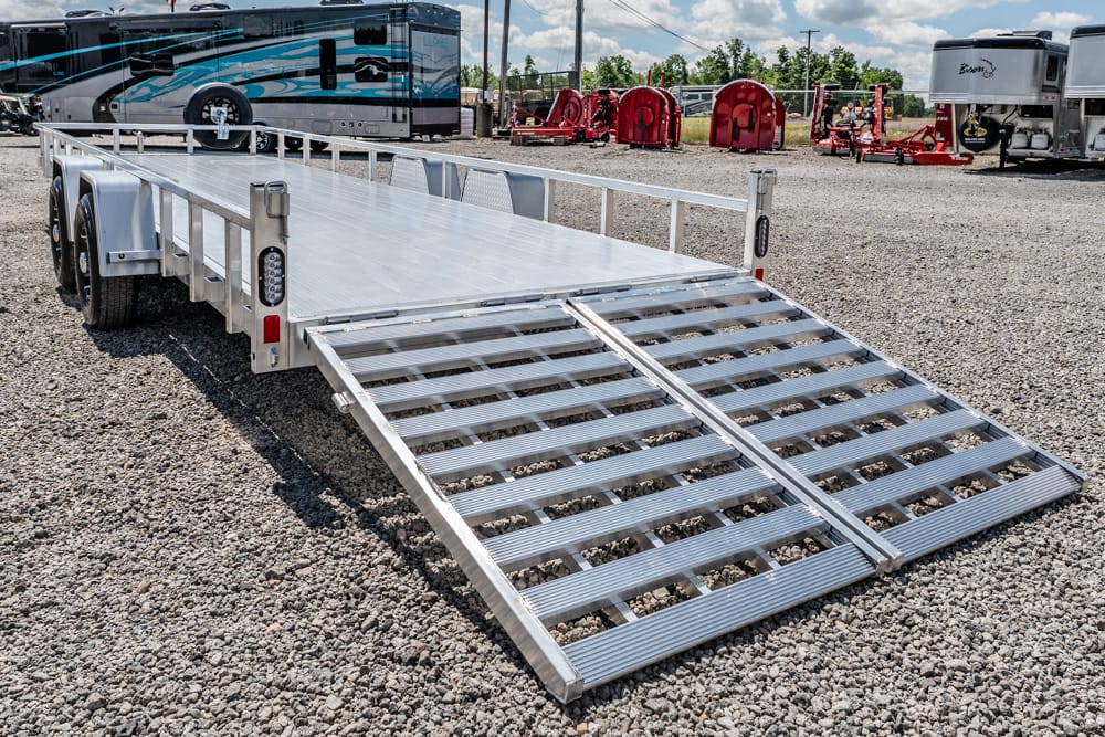 Aluminum flatbed trailer with dual loading ramps lowered to the ground on a gravel lot, with RVs and equipment in the background.