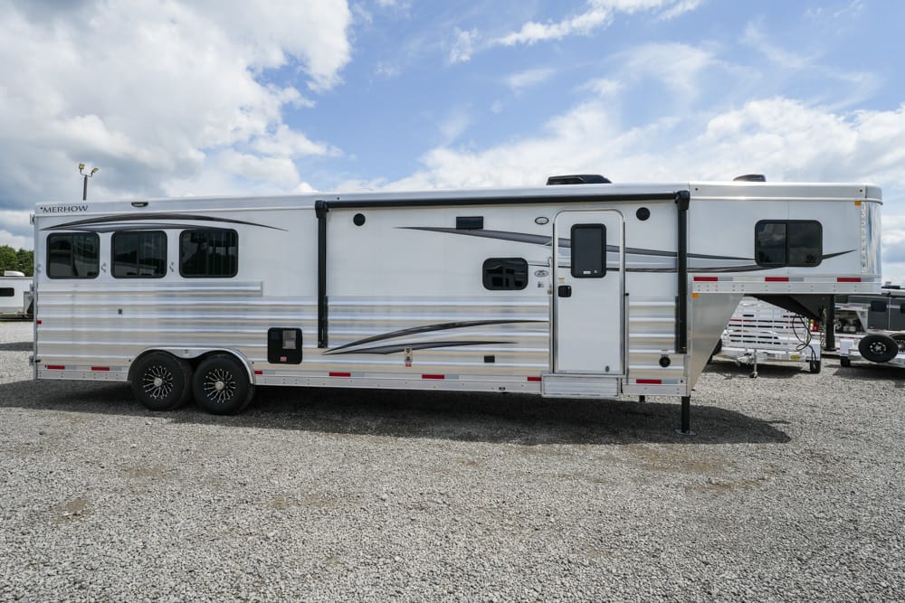 Side view of a white long travel trailer with decorative blue/gray stripes on a gravel lot, showing wheels, windows, door, and a raised hitch/overhang area at the front-right edge.