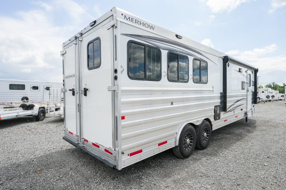 White Merhow aluminum horse trailer with two axles and multiple side windows parked on a gravel lot.
