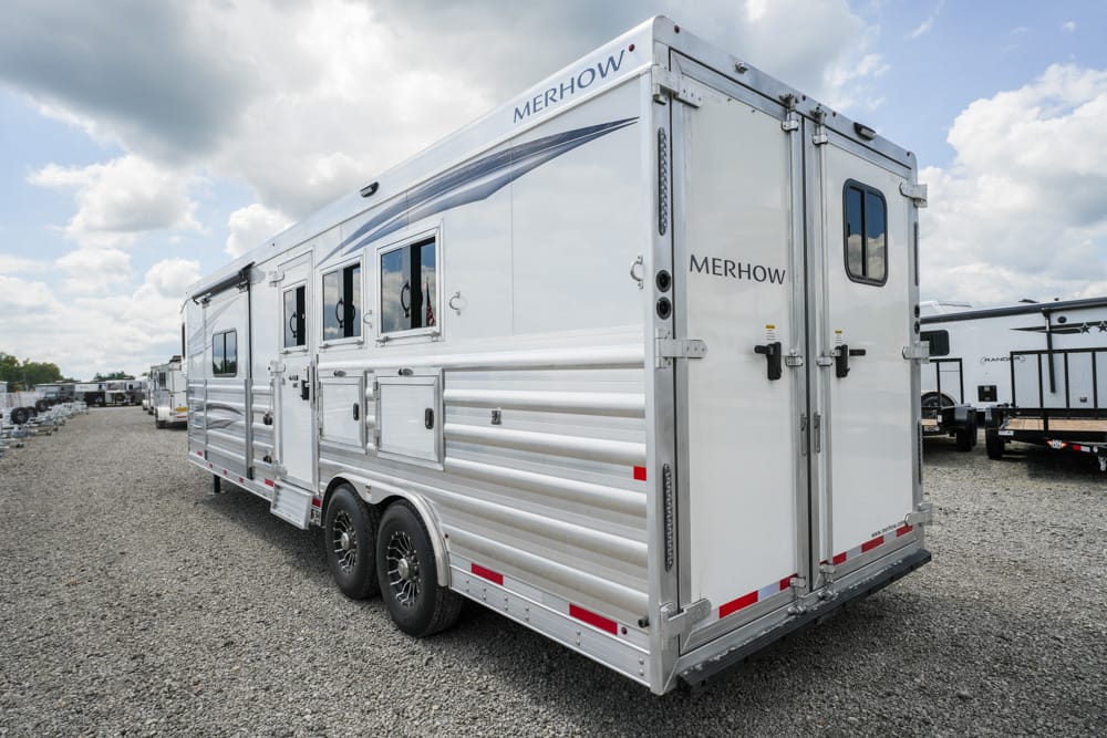 Silver Merhow horse trailer with three side windows and dual axles on a gravel lot in sunlight.