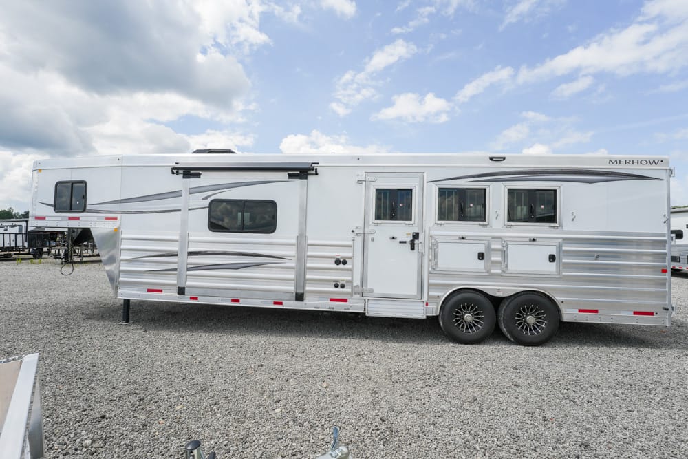 Long silver Merhow horse trailer with multiple windows and storage compartments on a gravel lot.