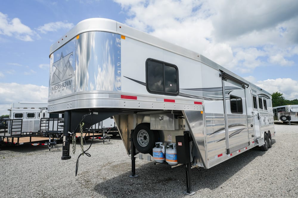 Silver gooseneck horse trailer with Merhow Summit branding, parked on a gravel lot with other trailers nearby.
