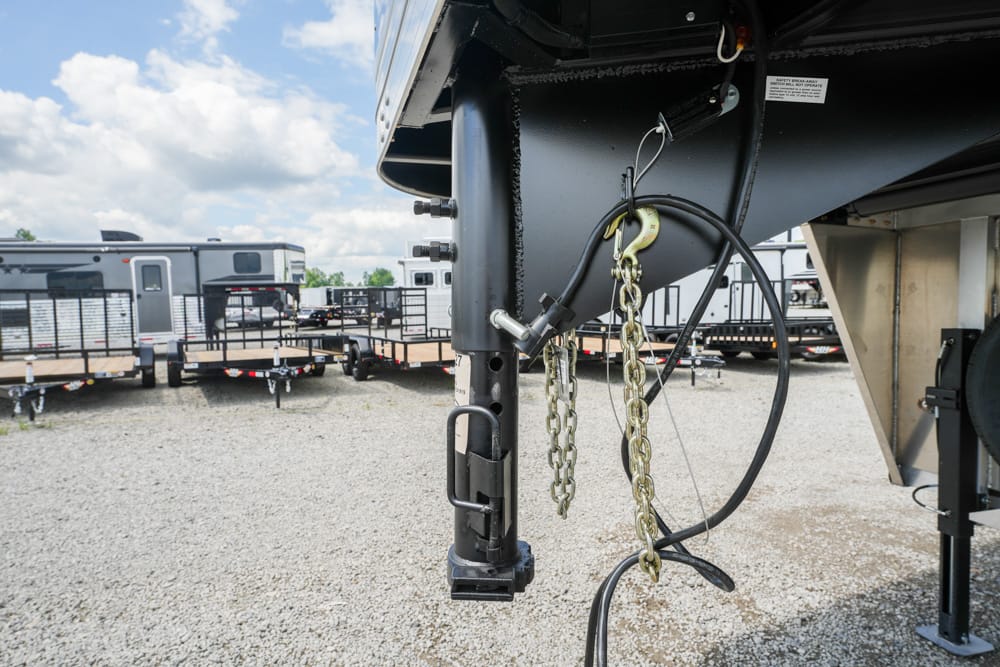 Close-up of a trailer hitch with safety chains and jack on a gravel lot; other trailers are visible in the background.
