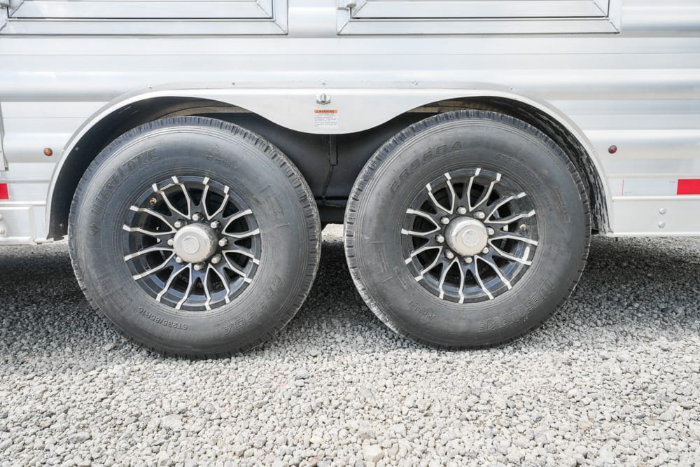 Dual-axle trailer with two black-rimmed tires on a gravel surface beneath a silver aluminum body.