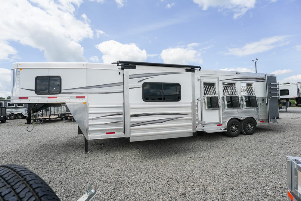 Long silver aluminum horse trailer with multiple windows, parked on a gravel lot under a blue sky.