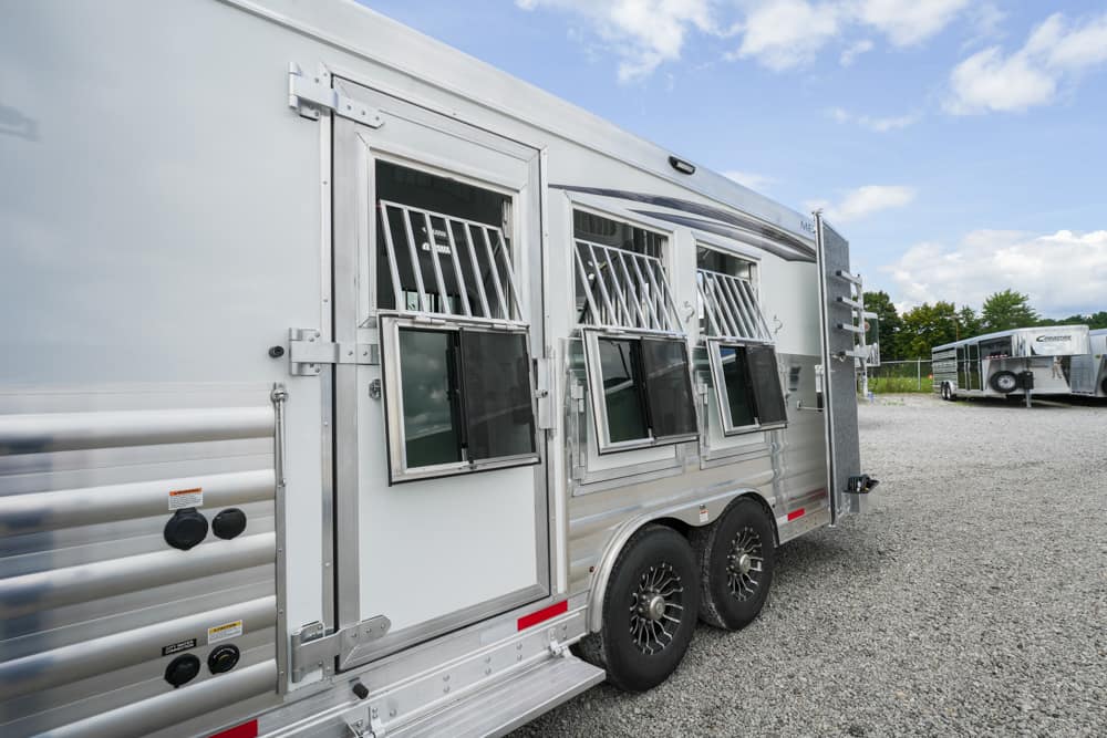Side view of a silver horse trailer with barred windows and dual axles, parked on a gravel lot; another trailer visible in the background