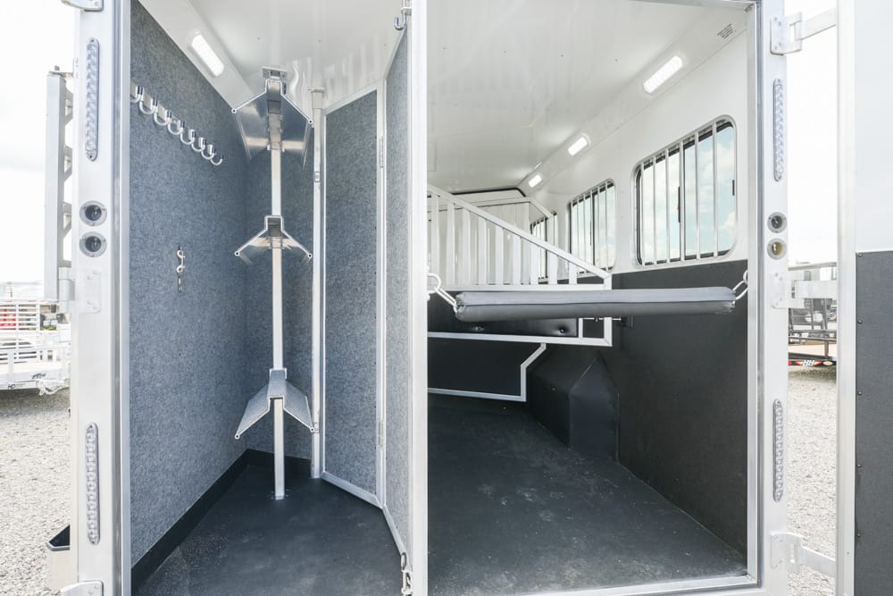 Interior of a horse trailer showing a tack/storage area with hooks and metal saddle racks next to a partition, and a separate guarded horse stall section with a padded lower wall and a wake-up bed area for transport.