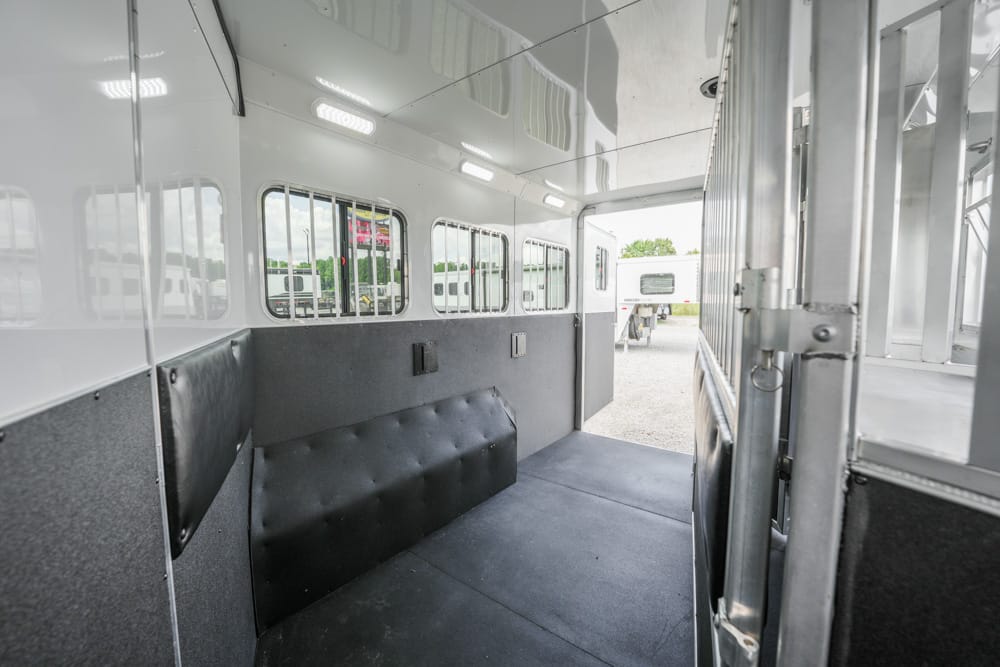 Interior of a white horse trailer with grey padded walls, bench seating along the left, and an open rear ramp revealing outside trailers