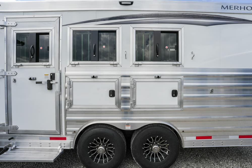 Silver horse trailer side view with two windows, storage compartments, and dual wheels visible on a gravel surface.