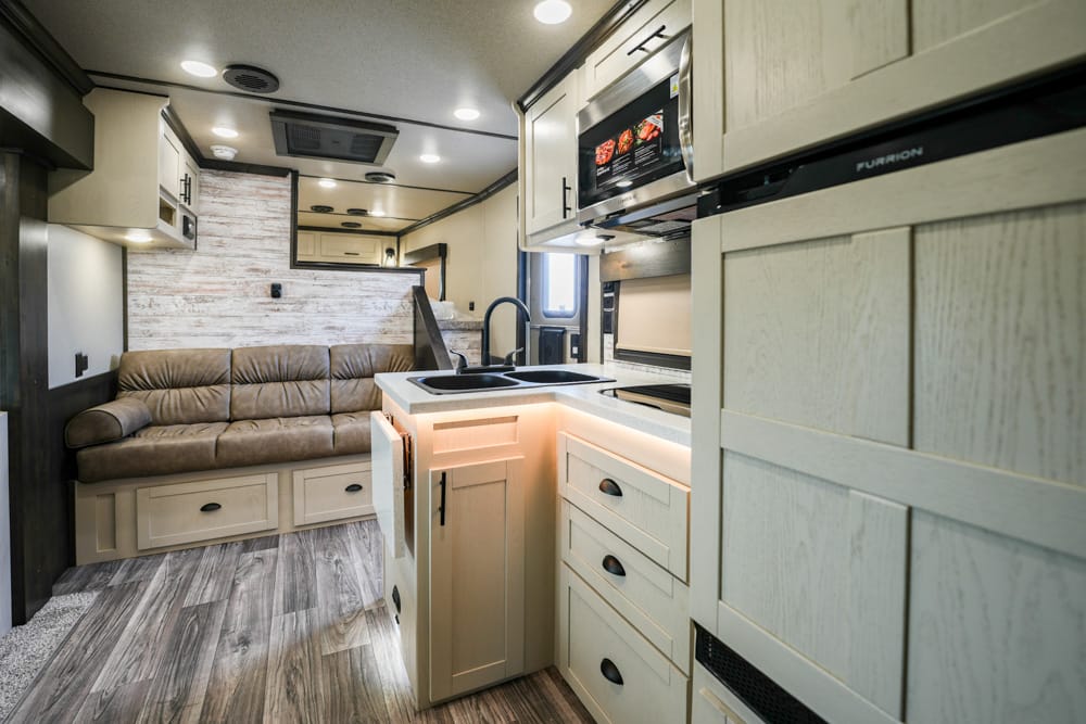 Interior of an RV kitchen and living area: beige couch along a brick-accent wall, cream cabinets, and a central island with sink and stove.
