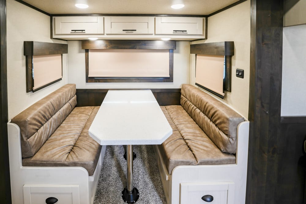 RV dining nook with beige cushioned benches, white central table, and overhead cabinets; windows with blinds above.