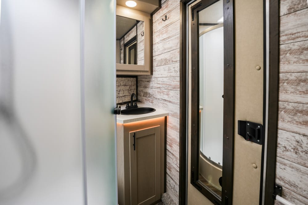 Compact RV bathroom with frosted glass shower door, sink, and beige cabinet under a mirror on weathered wood paneling