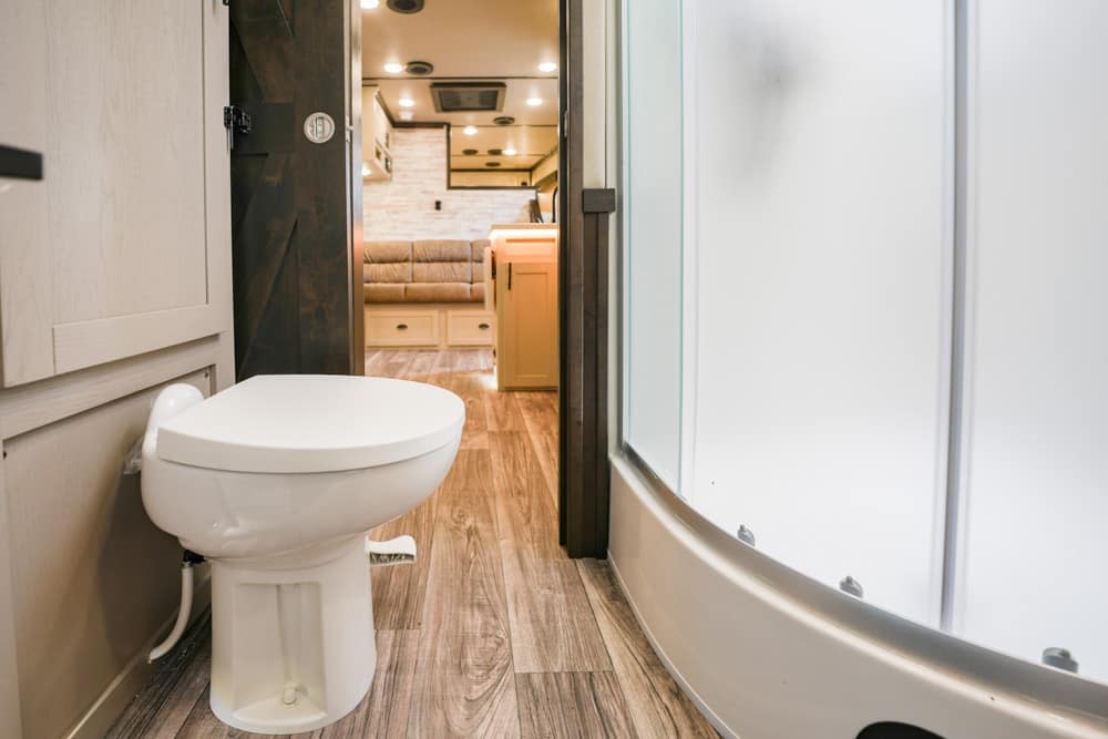 White toilet in an RV bathroom with a curved glass shower on the right and wood-look flooring; living area visible in the background.