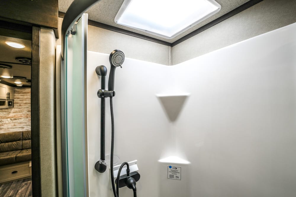 RV shower stall with black handheld showerhead on white wall and glass door on the left, ceiling light above