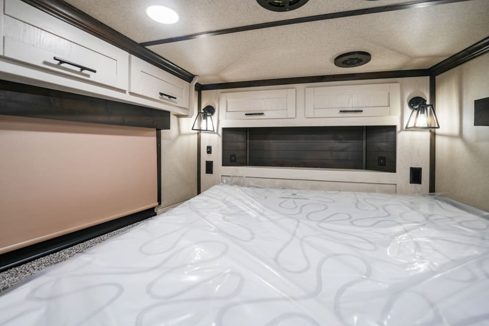 Cozy RV bedroom with a wide bed, white cabinets above, and black wood accents on the headboard wall