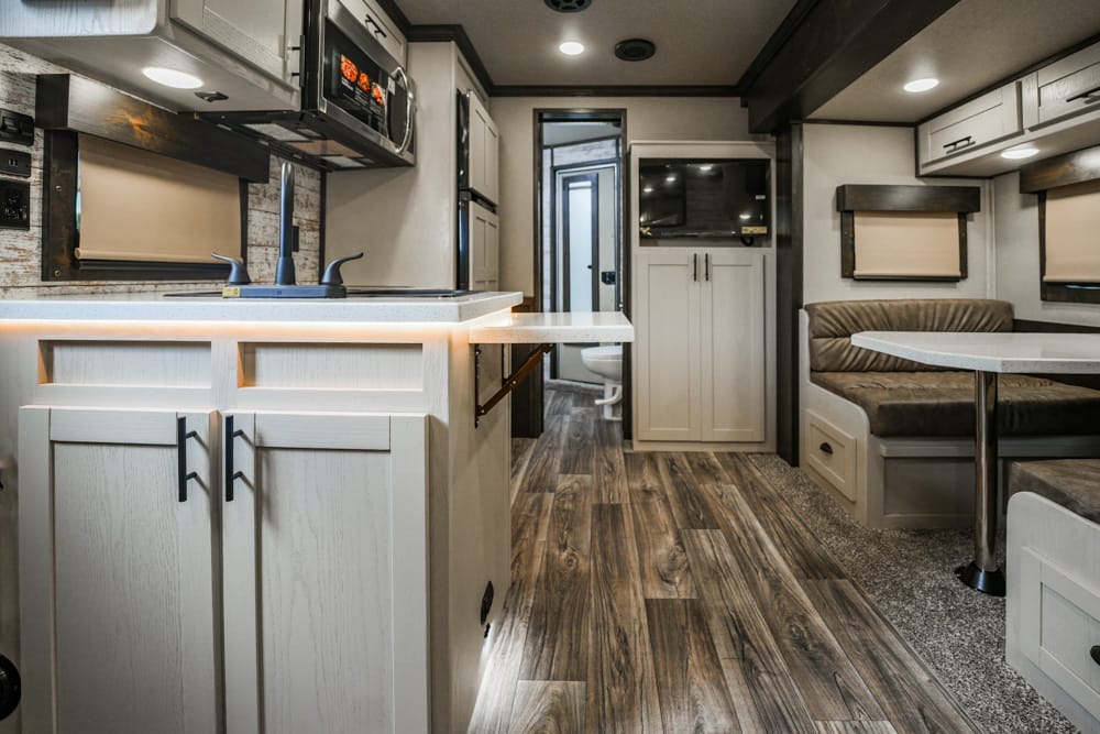 Bright RV interior with a white kitchen island, sink, and overhead microwave, next to a dinette booth with brown cushions.