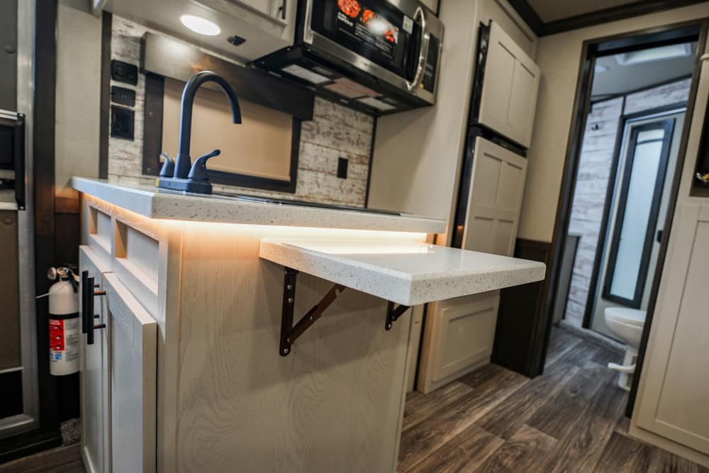 Interior of an RV kitchen: light wood island with a pull-out counter, black curved faucet, under-cabinet lighting, and a doorway to a compact bathroom.
