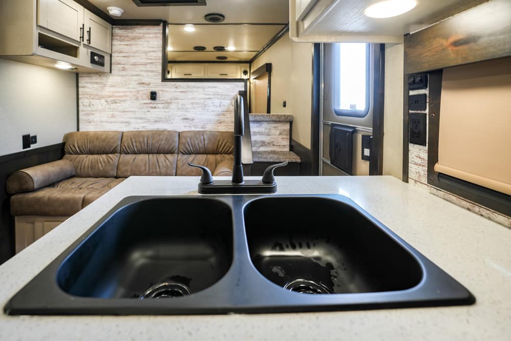 Double-basin sink with a modern faucet on a light speckled countertop in an RV kitchen, beige sofa and cabinets visible in the background.