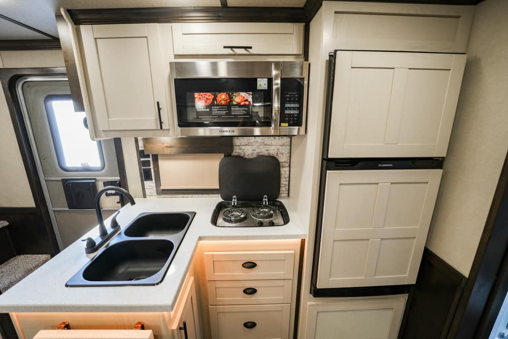 Compact RV kitchen with a double sink, two-burner stove, microwave above, and cream cabinetry.