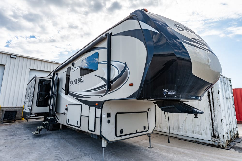 Sanibel fifth-wheel RV trailer parked on a concrete lot beside a storage building and shipping container.