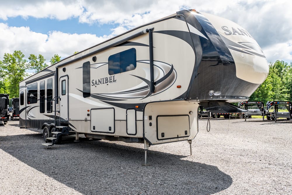 A large fifth-wheel RV trailer parked on a gravel lot, with 'SANIBEL' branding and decorative swirls on the side.
