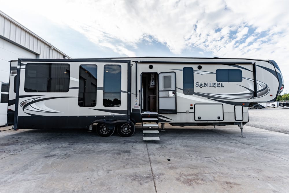 Large fifth-wheel RV trailer parked on a concrete lot, side view shows 'Sanibel' branding and decorative graphics.