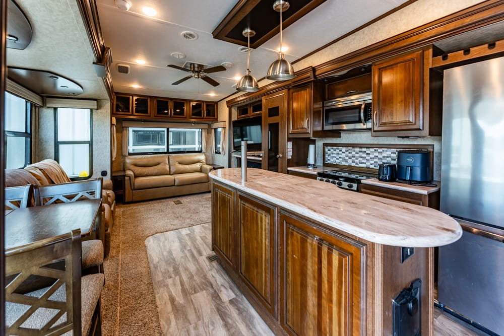Interior of a luxury RV kitchen with wooden cabinets, stove, fridge, and a marble island island; living area visible in background.