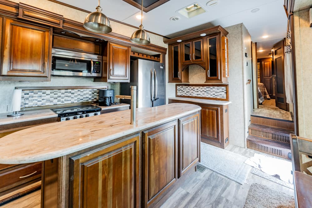 Wooden kitchen in a luxury motorhome with an island, stainless steel fridge, microwave, and pendant lights above the countertop.