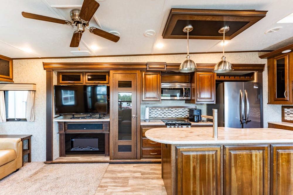Interior of a wooden RV kitchen with an island, stainless steel fridge, microwave, and overhead pendant lights; living area with a wall-mounted TV and fireplace.