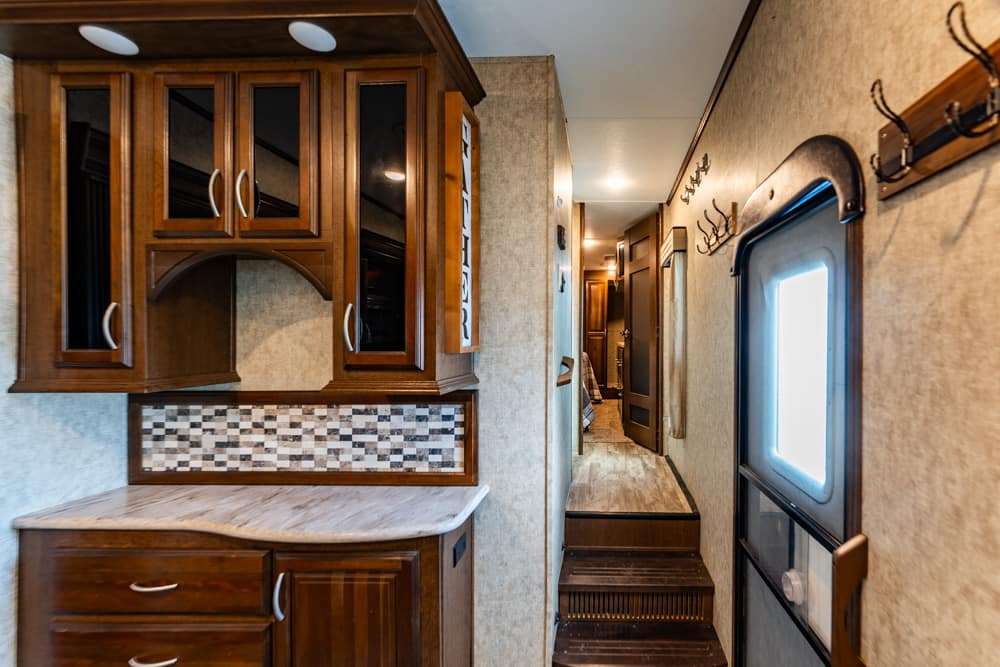 Interior of a recreational vehicle with dark wood kitchen cabinets, a curved countertop, and a mosaic tile backsplash; a narrow hallway leads to the back rooms.