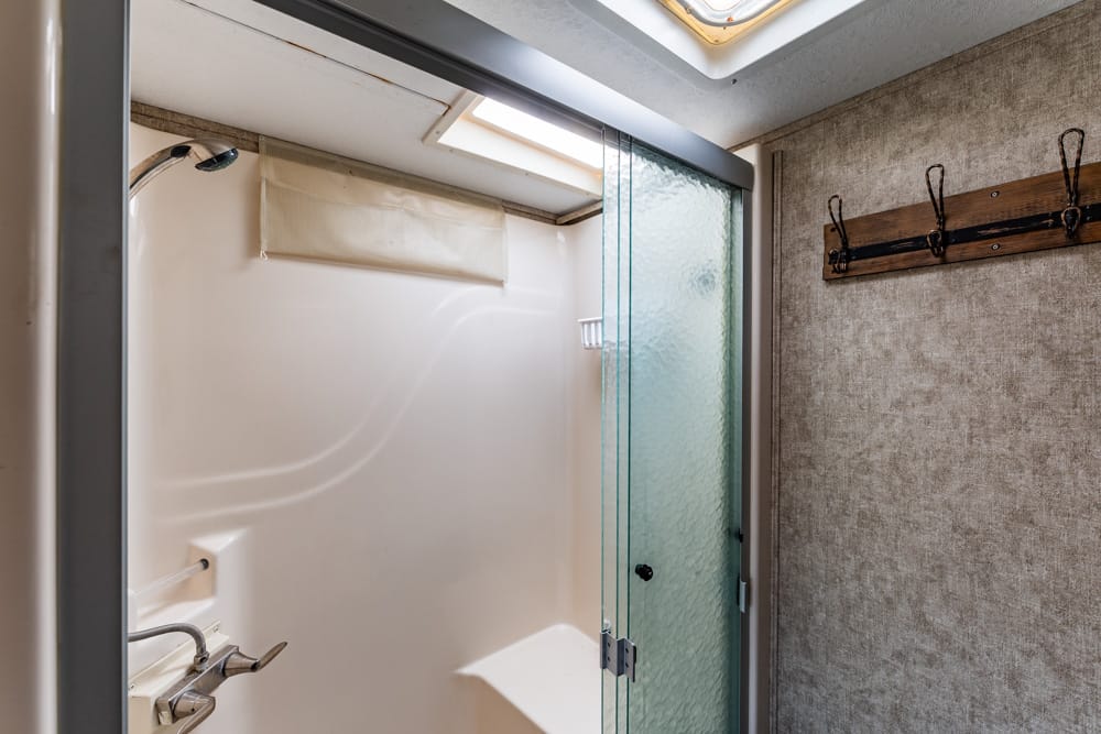 Shower stall with frosted glass door, white tub, and a skylight above in a small bathroom corner with beige walls and a wooden coat rack on the right wall