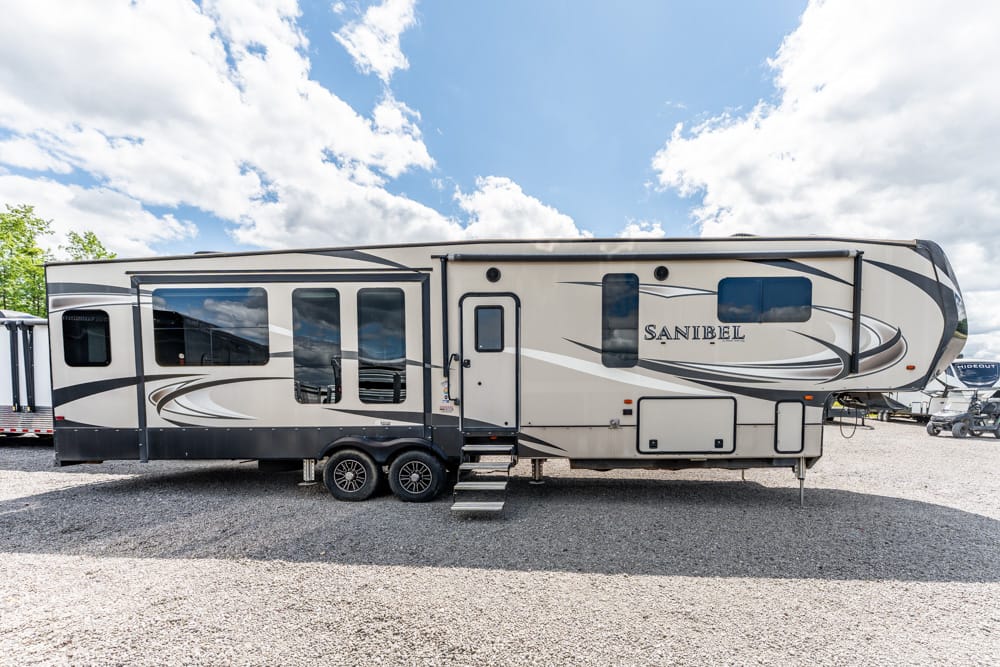 Side view of a beige and gray Sanibel fifth-wheel RV trailer parked on a gravel lot, with steps at the entry door and large windows.