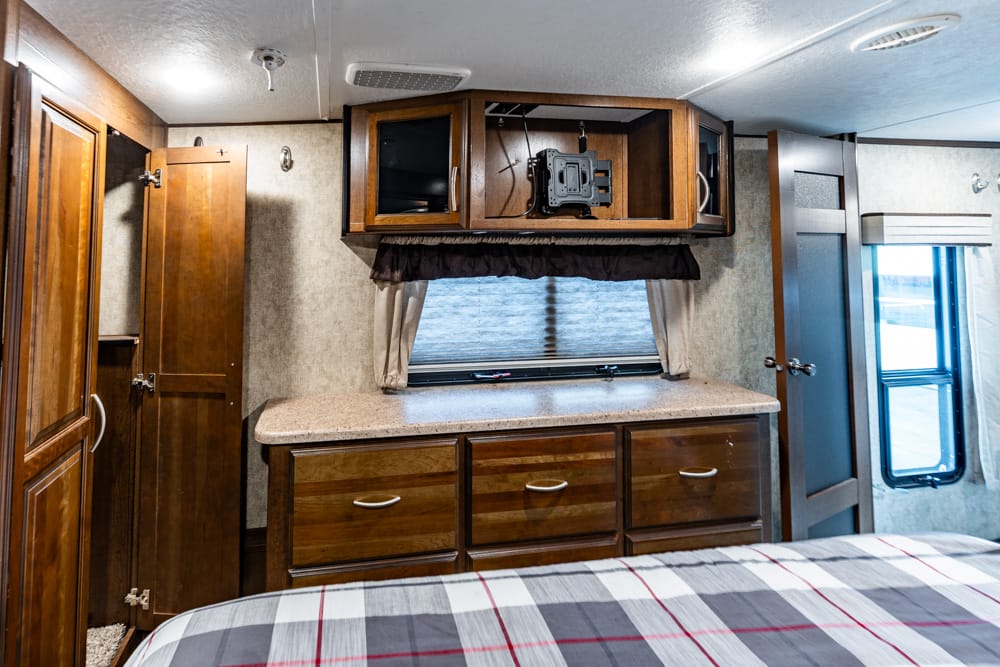 RV bedroom interior with dark wood cabinets, countertop, and a bed in the foreground; window with blinds behind the counter.