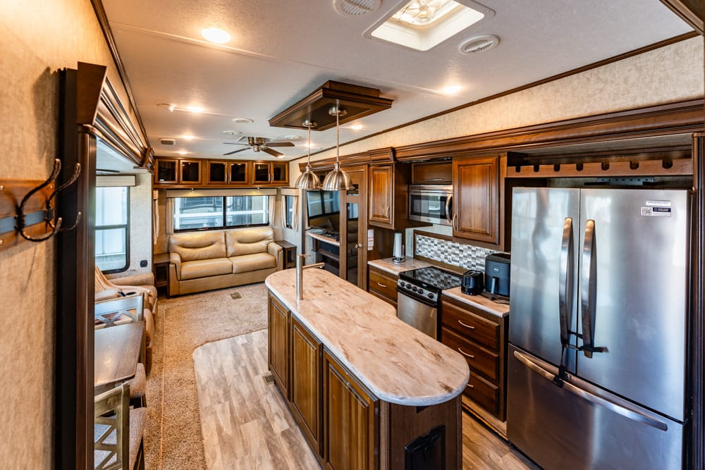 Interior view of a luxury RV kitchen and living area with a wooden island, stainless steel fridge, and beige sofa in the background.