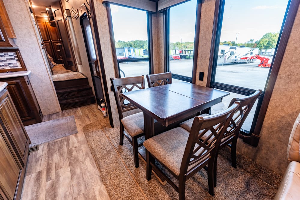 Interior of a recreational vehicle dining area with a dark wood table and four upholstered chairs, large windows overlooking parked trailers outside.
