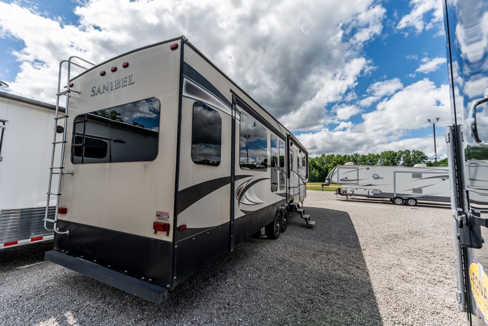Rear view of a beige and gray Sanibel travel trailer parked on a gravel lot, with other RVs and a cloudy blue sky in the background.
