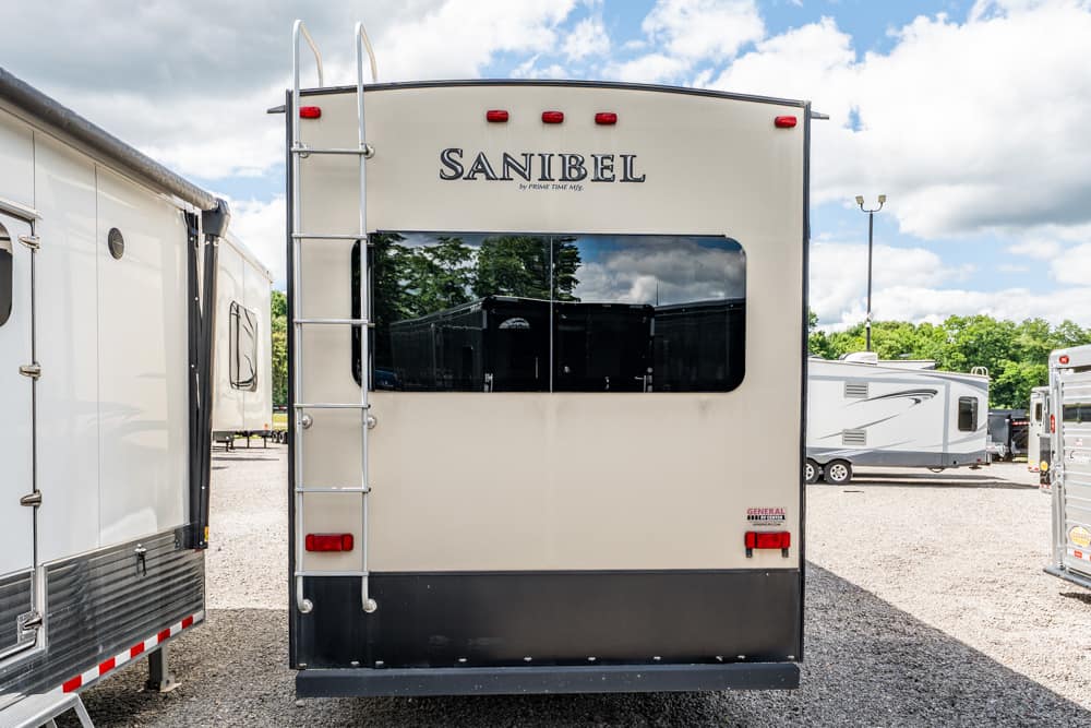 Rear view of a beige Sanibel travel trailer with a vertical ladder on the left, a large rear window, and red brake lights on a gravel RV lot.