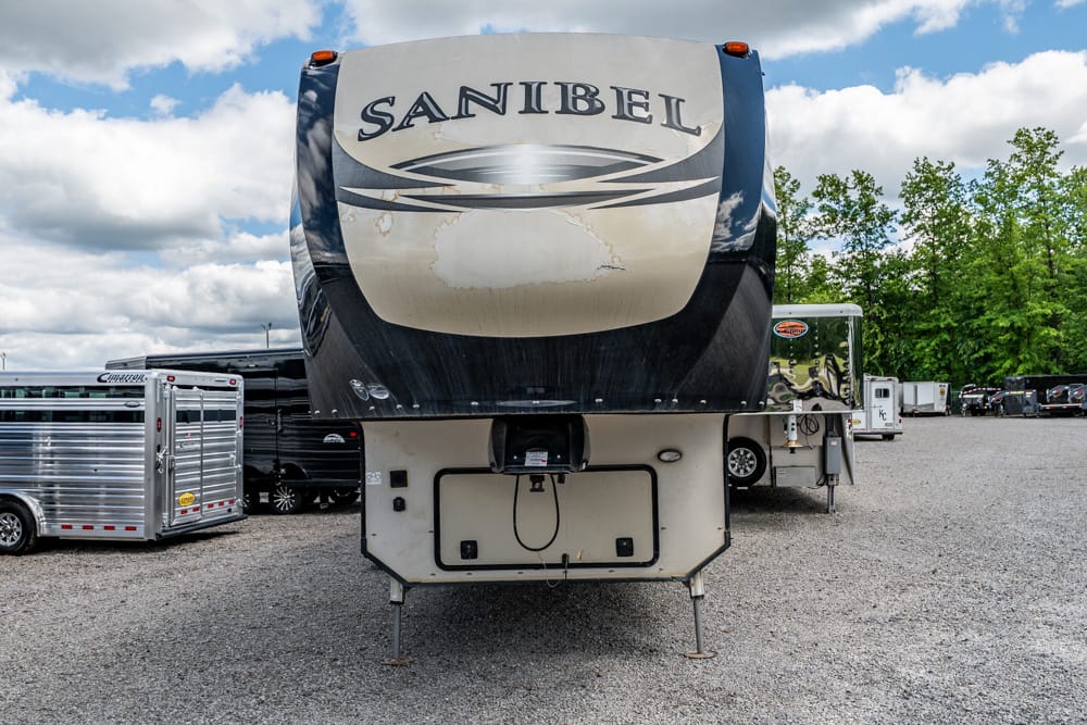 Front view of a large Sanibel fifth-wheel RV parked on a gravel lot, with other trailers and trees in the background.