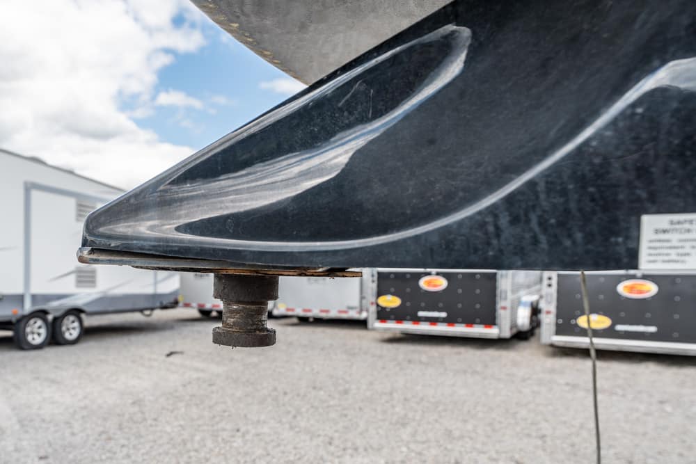 Close-up of a rusted trailer jack under the bow of a dark boat hull, with gravel and trailers in the background