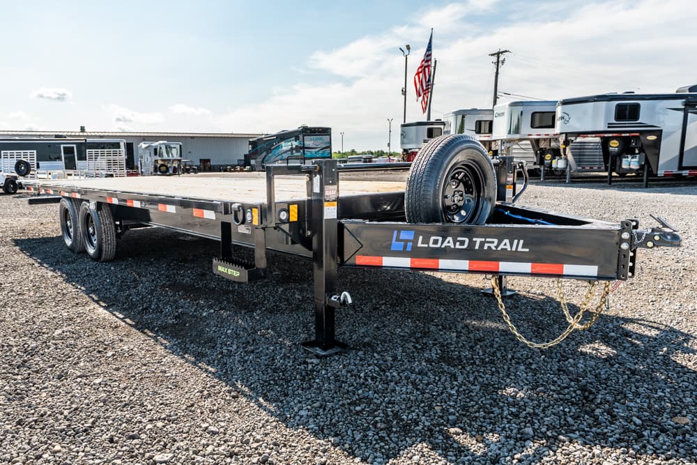 Load Trail flatbed trailer with spare tire and gooseneck hitch in a gravel lot near RVs and a flag in the background.