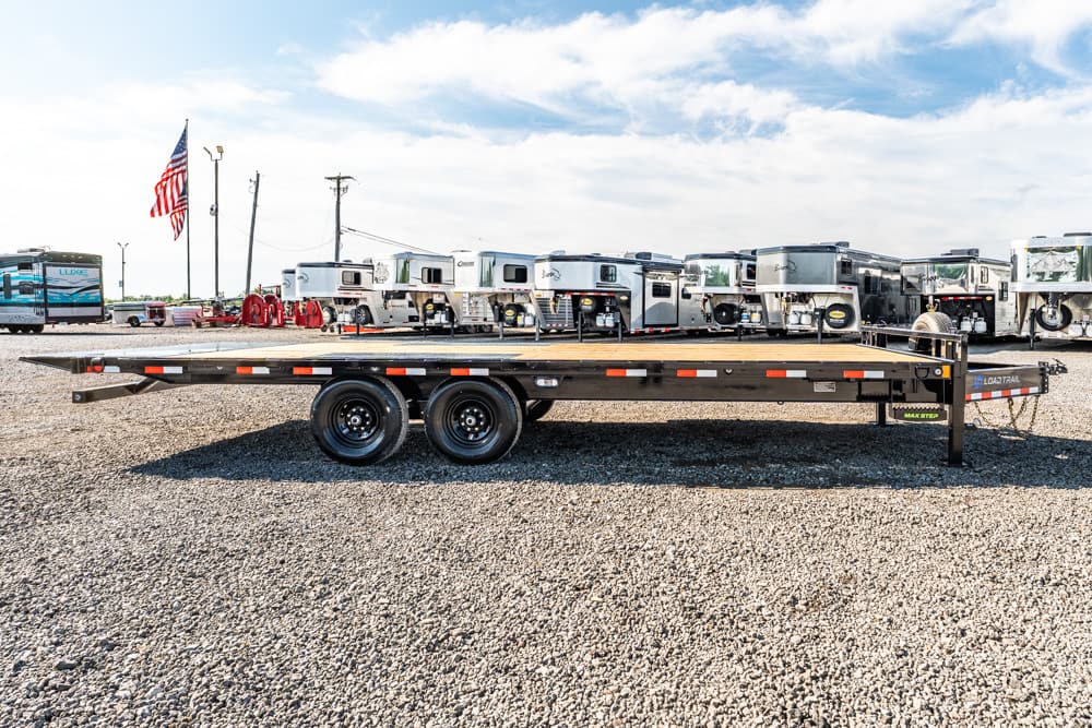 Flatbed trailer with dual axles parked on a gravel lot, row of RVs in the background and an American flag nearby.