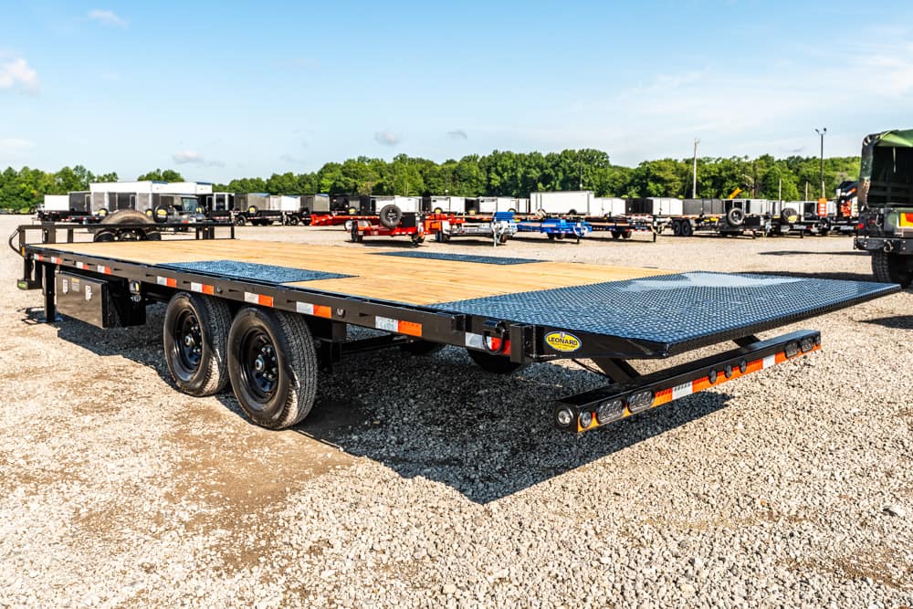 Black tandem-axle flatbed trailer with a wood deck and metal ramp, parked on a gravel lot with other trailers in the background.
