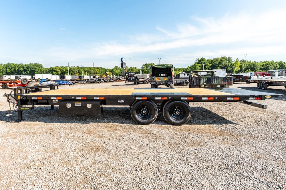 Black flatbed trailer with wooden deck, dual rear wheels, parked on gravel lot with other trailers in the background reading distancewise.