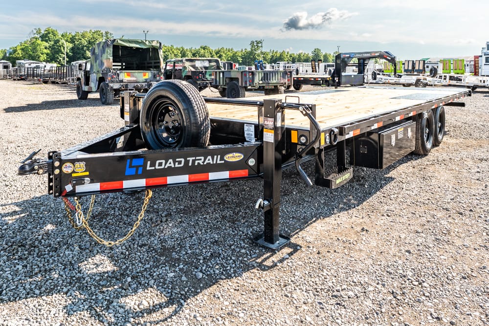 Flatbed trailer with a spare tire on the tongue, in a gravel lot with trucks in the background; labeled 'Load Trail' for hauling equipment.