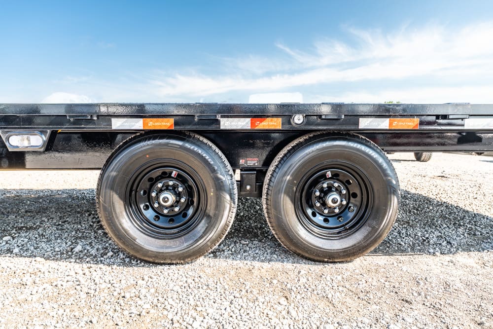 Dual-axle trailer wheels with black rims on a flatbed trailer parked on gravel under a blue sky.