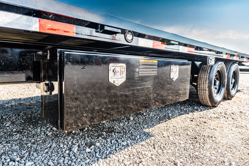 Black metal storage box mounted under a flatbed trailer on a gravel lot, featuring two latches and a warning label.
