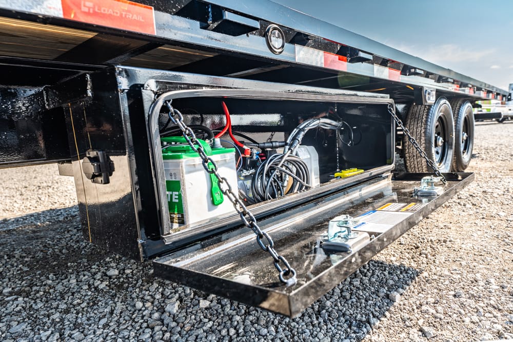 Open storage compartment on a flatbed trailer showing a battery, cables, and hoses inside on gravel ground.