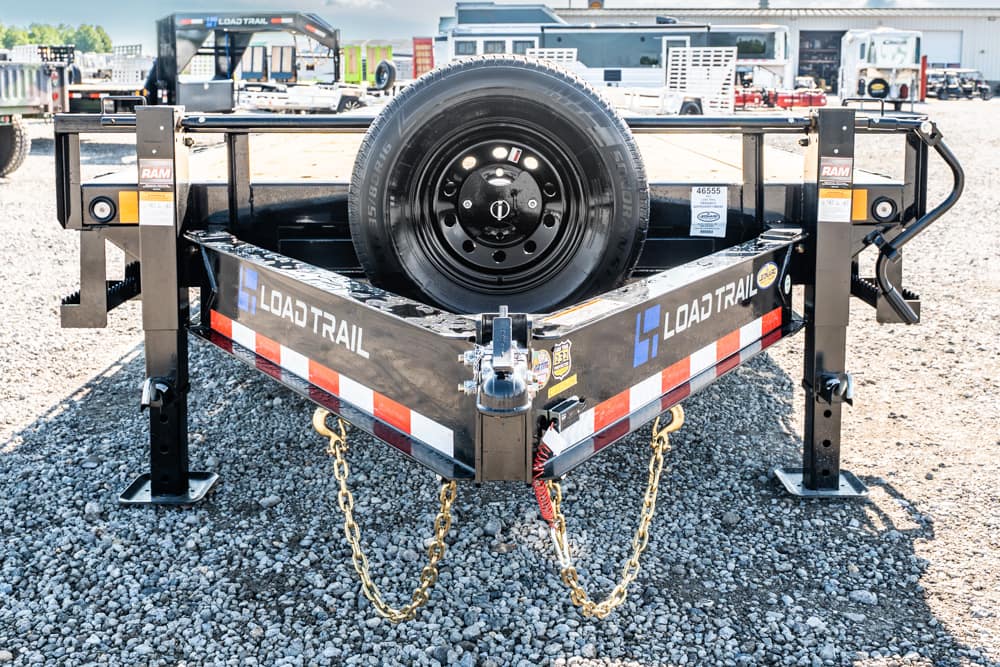 Black Load Trail trailer frame with a mounted spare tire facing the viewer on a gravel lot; safety chains hang from the hitch area.