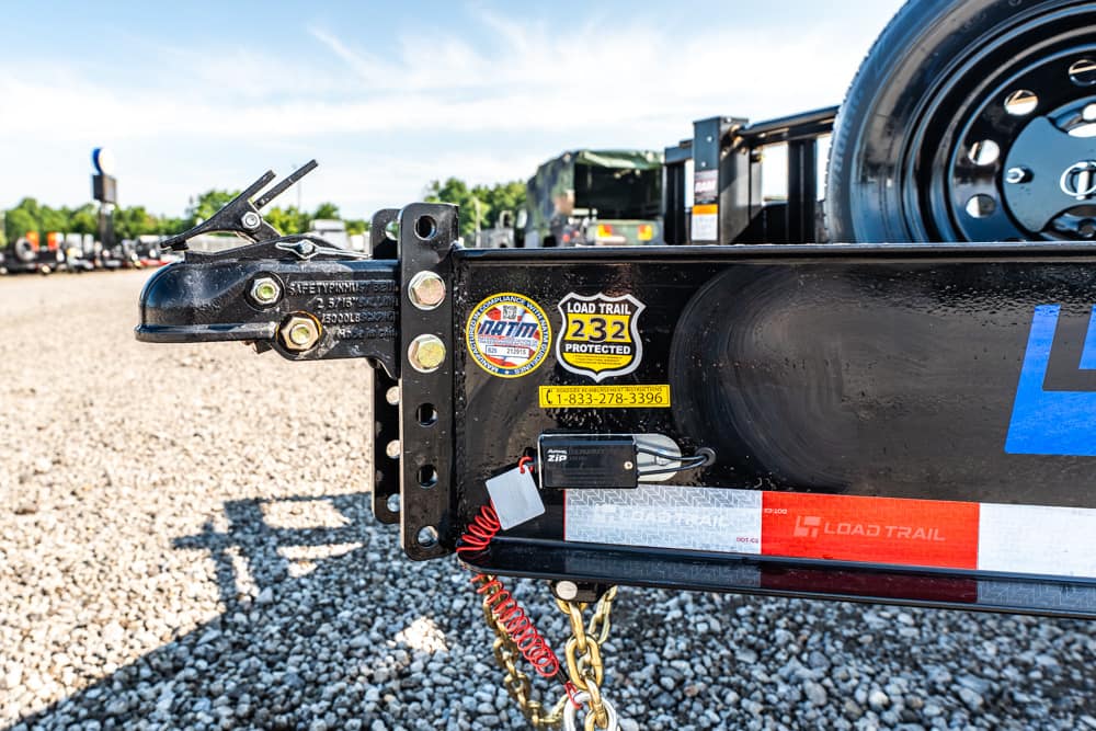 Black trailer hitch with safety latch and bolt-on components on a gravel lot; spare wheel attached to the trailer frame in the background.