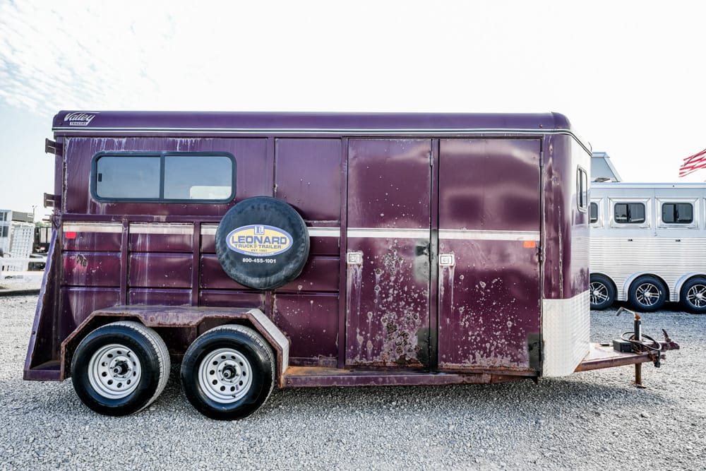 Purple horse trailer with dual axles and spare tire mounted on side, parked on a gravel lot with another trailer in the background.